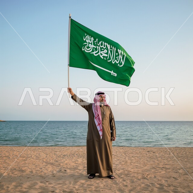 The concept of pride and honor for the homeland, celebrating national holidays and occasions, remembering Saudi National Day September 23, commemorating Flag Day March 11, a Saudi Gulf Arab man wearing the traditional dress and shemagh stands on the beach and raises the flag of the Kingdom of Saudi Arabia in his hand