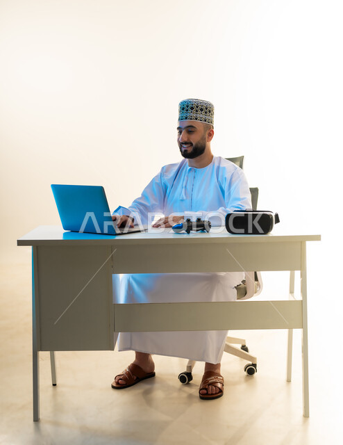 Using modern technical devices, simulating the imaginary world, a portrait of a young Gulf Arab Omani man wearing a dishdasha and a kuma sitting in front of a desk working on a computer, next to him are VR glasses and a joystick, integrating technology into work