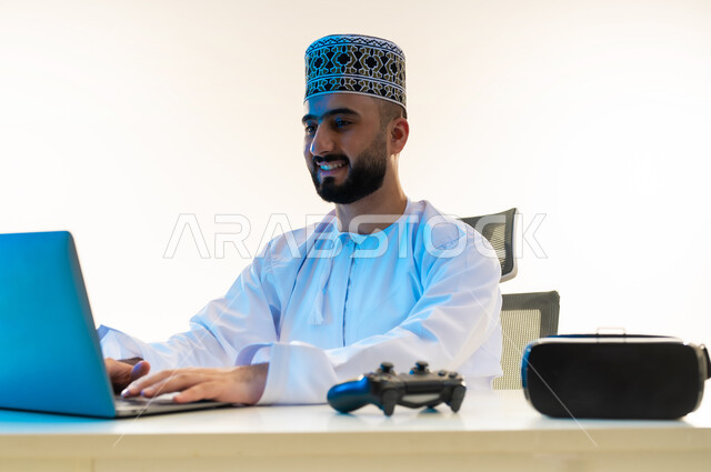 دمج التكتولوجيا بالعمل، استخدام الأجهزة التقنية الحديثة, محاكاة العالم الخيالي، بورتريه مقرب لشاب عربي خليجي عماني يرتدي الدشداشة والكمة يجلس امام المكتب يعمل على جهاز الحاسوب بجانبه نظارة الفي ار وعصا التحكم