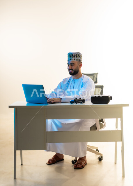 استخدام الأجهزة التقنية الحديثة, محاكاة العالم الخيالي، بورتريه لشاب عربي خليجي عماني يرتدي الدشداشة والكمة يجلس امام المكتب يعمل على جهاز الحاسوب بجانبه نظارة الفي ار وعصا التحكم، دمج التكتولوجيا بالعمل