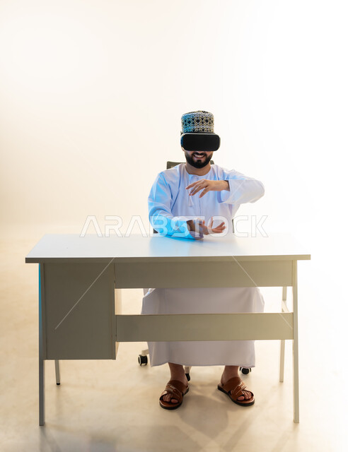محاكاة العالم الخيالي، التطور التقني والتكنولوجي، المتعة بالمشاهدة عبر نظارة الواقع الافتراضي، بورتريه لشاب عربي خليجي عماني يرتدي الدشداشة والكمه يجلس امام المكتب ينظر من خلال نظارات الفي ار يرفع يديه بايماءات حمل عنصر هولوجرامي، خلفية بيضاء