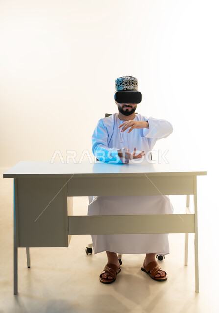 محاكاة العالم الخيالي، التطور التقني والتكنولوجي، المتعة بالمشاهدة عبر نظارة الواقع الافتراضي، بورتريه لشاب عربي خليجي عماني يرتدي الدشداشة والكمه يجلس امام المكتب ينظر من خلال نظارات الفي ار يرفع يديه بايماءات حمل عنصر هولوجرامي، خلفية بيضاء
