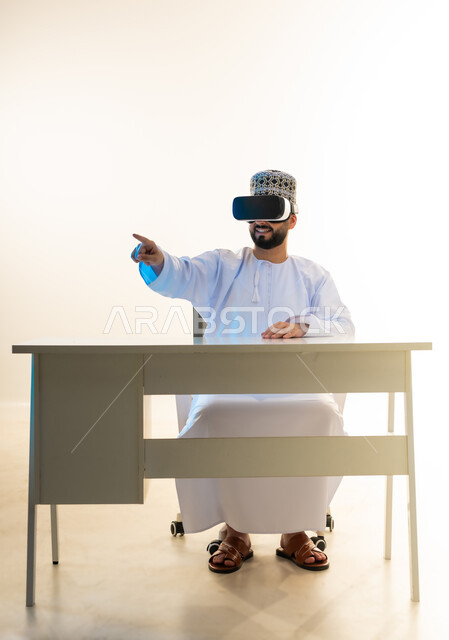 تحديد موقع شيء ما، التطور التقني والتكنولوجي، المتعة بالمشاهدة عبر نظارة الواقع الافتراضي، بورتريه لشاب عربي خليجي عماني يرتدي الدشداشة والكمه يجلس امام المكتب ينظر من خلال نظارات الفي ار يشير باصبع السبابة لمكان ما، محاكاة العالم الخيالي، خلفية بيضاء
