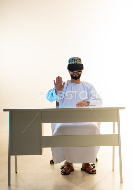 تعابير الامتناع عن شيء ما، التطور التقني والتكنولوجي، المتعة بالمشاهدة عبر نظارة الواقع الافتراضي، بورتريه لشاب عربي خليجي عماني يرتدي الدشداشة والكمه يجلس امام المكتب ينظر من خلال نظارات الفي ار يرفع يده بايماءات الرفض، محاكاة العالم الخيالي، خلفية بيضاء