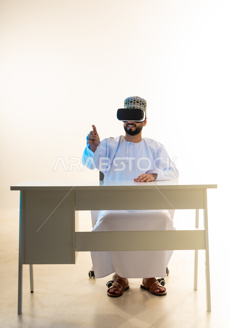 Locating something, technical and technological development, enjoy watching through virtual reality glasses, portrait of a young Arab Gulf Omani man wearing a dishdasha and a cloak sitting in front of a desk looking through VR glasses pointing with his index finger to a place, simulating the imaginary world, white background
