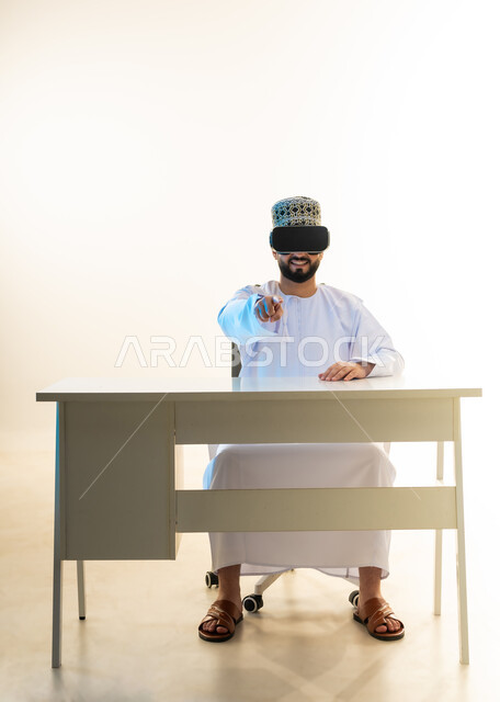 تحديد موقع شيء ما، التطور التقني والتكنولوجي، المتعة بالمشاهدة عبر نظارة الواقع الافتراضي، بورتريه لشاب عربي خليجي عماني يرتدي الدشداشة والكمه يجلس امام المكتب ينظر من خلال نظارات الفي ار يشير باصبع السبابة الكاميرا، محاكاة العالم الخيالي، خلفية بيضاء