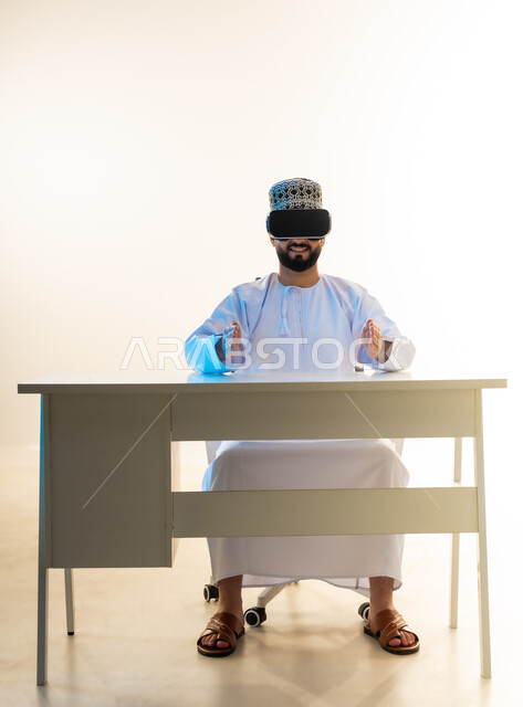 محاكاة العالم الخيالي، المتعة بالمشاهدة عبر نظارة الواقع الافتراضي، التطور التقني والتكنولوجي، بورتريه لشاب عربي خليجي عماني يرتدي الدشداشة والكمه يجلس امام المكتب ينظر من خلال نظارات الفي ار يرفع يديه بايماءات حمل عنصر ما، خلفية بيضاء