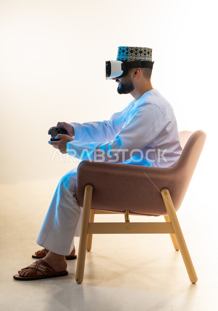 Interaction and integration with video games, simulation of the imaginary world, modern entertainment electronic devices and technologies, spending a fun and entertaining time, side portrait of an Arab Gulf Omani man wearing a dishdasha, a turban and virtual reality glasses sitting on a comfortable chair holding a joystick, white background