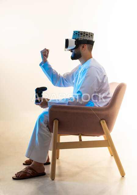 ايماءات الفوز والنجاح، محاكاة العالم الخيالي، قضاء التفاعل والاندماج مع ألعاب الفيديو، قضاء وقت ممتع ومسلي، بورتريه من الجانب لرجل عربي خليجي عماني يرتدي الدشداشة والكمة ونظارة الواقع الافتراضي يجلس على كرسي مريح يحمل عصا التحكم يرفع يده بتعابير النصر، خلفية بيضاء