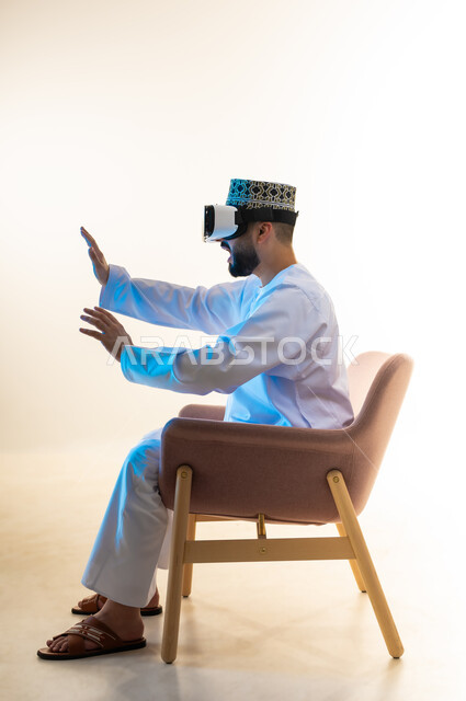 Technical and technological development, fun and entertainment watching through virtual reality glasses, side portrait of a young Gulf Arab Omani man wearing a dishdasha and a kimono sitting on a comfortable chair looking through VR glasses raising his hands with gestures of integration, simulating the imaginary world, white background