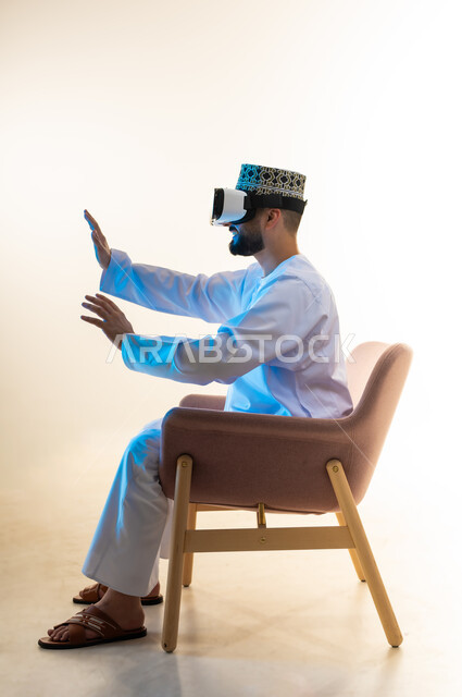المتعة والتسلية بالمشاهدة عبر نظارة الواقع الافتراضي، التطور التقني والتكنولوجي, بورتريه من الجانب لشاب عربي خليجي عماني يرتدي الدشداشة والكمه يجلس على كرسي مريح ينظر من خلال نظارات الفي ار يرفع يديه بايماءات الاندماج، محاكاة العالم الخيالي، خلفية بيضاء