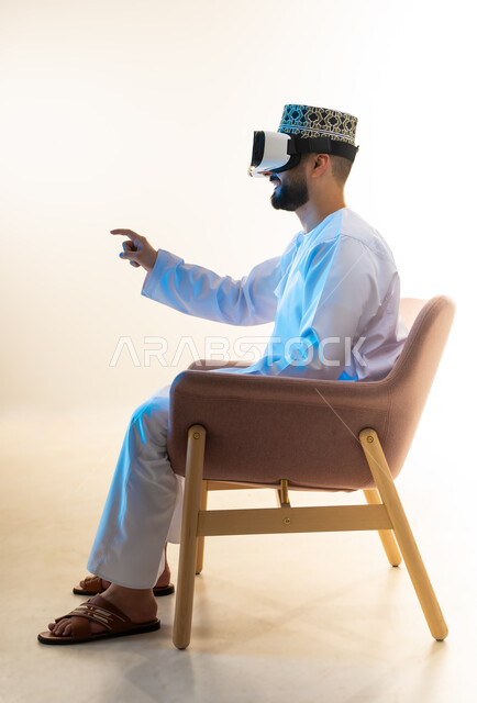 التطور التقني والتكنولوجي, المتعة والتسلية بالمشاهدة عبر نظارة الواقع الافتراضي، بورتريه من الجانب لشاب عربي خليجي عماني يرتدي الدشداشة والكمه يجلس على كرسي مريح ينظر من خلال نظارات الفي ار يشير باصبع السبابة لمكان ما، محاكاة العالم الخيالي، خلفية بيضاء