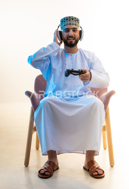 الاندماج والتفاعل مع ألعاب الفيديو، مفهوم الاصغاء والاستماع عبر الهيدفون، اجهزة وتقنيات الكترونية ترفيهيه حديثة، قضاء وقت ممتع ومسلي، بورتريه لرجل عربي خليجي عماني يرتدي الدشداشة والكمة وسماعة الرأس يجلس على كرسي مريح يحمل عصا التحكم، خلفية بيضاء