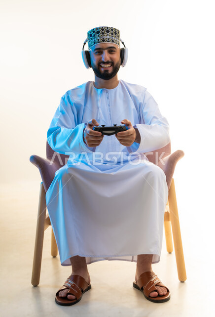 مفهوم الاصغاء والاستماع عبر الهيدفون، الاندماج والتفاعل مع ألعاب الفيديو، اجهزة وتقنيات الكترونية ترفيهيه حديثة، قضاء وقت ممتع ومسلي، بورتريه لرجل عربي خليجي عماني يرتدي الدشداشة والكمة وسماعة الرأس يجلس على كرسي مريح يحمل عصا التحكم، خلفية بيضاء