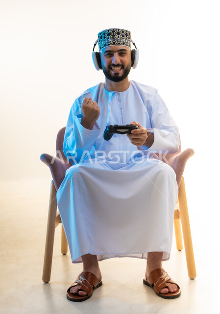 ايماءات الفوز والنجاح، مفهوم الاصغاء والاستماع عبر الهيدفون، اجهزة وتقنيات ترفيهيه حديثة، التفاعل مع ألعاب الفيديو، بورتريه لرجل عربي خليجي عماني يرتدي الدشداشة والكمة وسماعات الرأس يجلس على كرسي مريح يحمل عصا التحكم يرفع قبضة يده بتعابير النصر، خلفية بيضاء