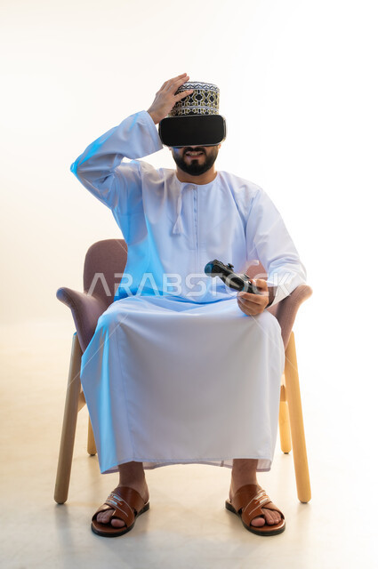 Simulating the imaginary world, spending interaction and integration with video games, spending a fun and entertaining time, portrait of an Arab Gulf Omani man wearing a dishdasha, a turban, and virtual reality glasses, sitting on a comfortable chair holding a joystick, putting his hand on his head with an expression of disappointment, white background