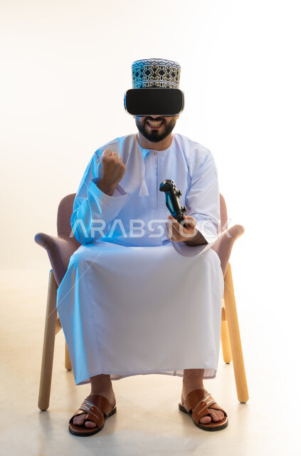 ايماءات الفوز والنجاح، محاكاة العالم الخيالي، قضاء التفاعل والاندماج مع ألعاب الفيديو، قضاء وقت ممتع ومسلي، بورتريه لرجل عربي خليجي عماني يرتدي الدشداشة والكمة ونظارة الواقع الافتراضي يجلس على كرسي مريح يحمل عصا التحكم يرفع يده بتعابير النصر، خلفية بيضاء