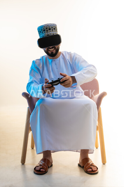 محاكاة العالم الخيالي، اجهزة وتقنيات الكترونية ترفيهيه حديثة, التفاعل والاندماج مع ألعاب الفيديو، قضاء وقت ممتع ومسلي، بورتريه لرجل عربي خليجي عماني يرتدي الدشداشة والكمة ونظارة الواقع الافتراضي يجلس على كرسي مريح يحمل عصا التحكم، خلفية بيضاء