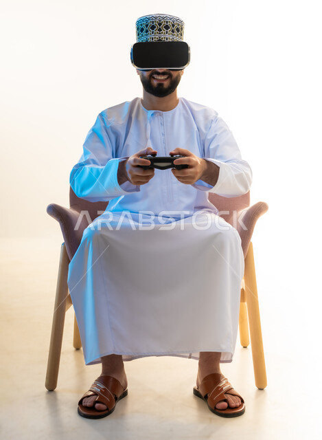 Modern entertainment electronic devices and technologies, simulating the imaginary world, interacting and merging with video games, spending a fun and entertaining time, portrait of an Arab Gulf Omani man wearing a dishdasha, a kimono and virtual reality glasses sitting on a comfortable chair holding a joystick, white background