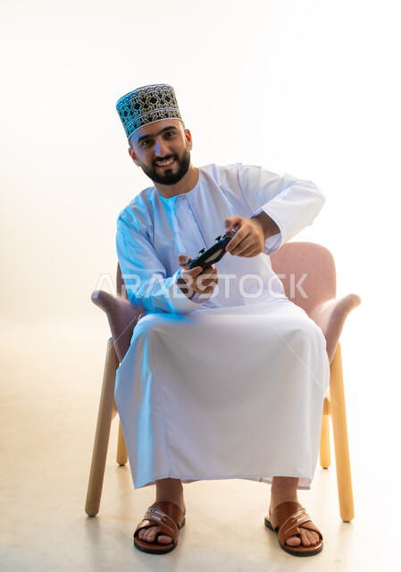 النظر الى الكاميرا بايماءات السرور، التفاعل والاندماج مع ألعاب الفيديو، قضاء وقت ممتع ومسلي، اجهزة وتقنيات الكترونية ترفيهيه حديثة, بورتريه لشاب عربي خليجي عماني يرتدي الدشداشة والكمة يجلس على كرسي مريح يحمل عصا التحكم، خلفية بيضاء