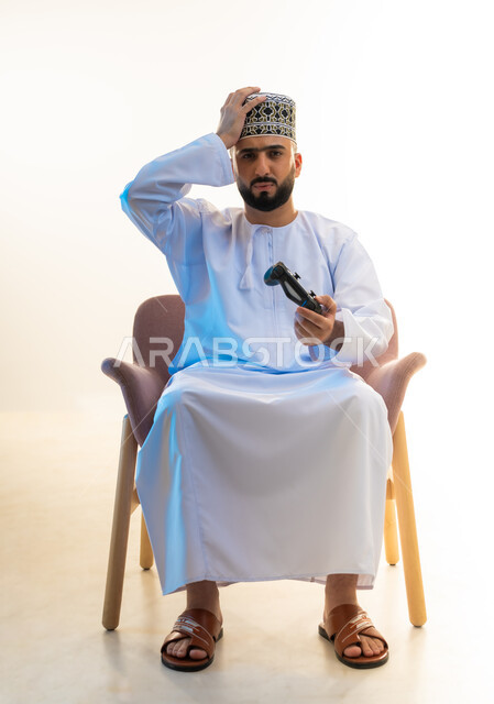 Interacting and merging with video games, having a fun and entertaining time, modern entertainment electronic devices and technologies, portrait of a young Gulf Arab Omani man wearing a dishdasha and a turban, sitting on a comfortable chair holding a joystick, putting his hand on his head with gestures of disappointment, white background