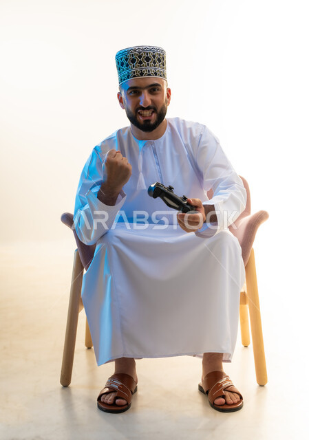 ايماءات الفوز والنجاح، التفاعل والاندماج مع ألعاب الفيديو، قضاء وقت ممتع ومسلي، اجهزة وتقنيات الكترونية ترفيهيه حديثة, بورتريه لشاب عربي خليجي عماني يرتدي الدشداشة والكمة يجلس على كرسي مريح يحمل عصا التحكم يرفع يده بتعابير النصر، خلفية بيضاء