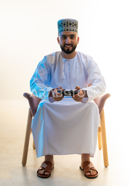 النظر الى الكاميرا بايماءات السرور، التفاعل والاندماج مع ألعاب الفيديو، قضاء وقت ممتع ومسلي، اجهزة وتقنيات الكترونية ترفيهيه حديثة, بورتريه لشاب عربي خليجي عماني يرتدي الدشداشة والكمة يجلس على كرسي مريح يحمل عصا التحكم، خلفية بيضاء