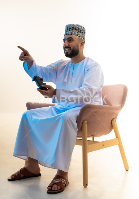 Locating a specific thing, interacting and merging with video games, having a fun and entertaining time, modern entertainment electronic devices and technologies, portrait of a young Arab Gulf Omani man wearing a dishdasha and a turban sitting on a comfortable chair holding a joystick pointing with his index finger to a place, white background