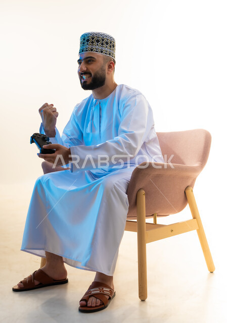 اجهزة وتقنيات الكترونية ترفيهيه حديثة, ايماءات الفوز والنجاح، التفاعل والاندماج مع ألعاب الفيديو، قضاء وقت ممتع ومسلي، بورتريه من الجانب لشاب عربي خليجي عماني يرتدي الدشداشة والكمة يجلس على كرسي مريح يحمل عصا التحكم يرفع يده بتعابير النصر، خلفية بيضاء