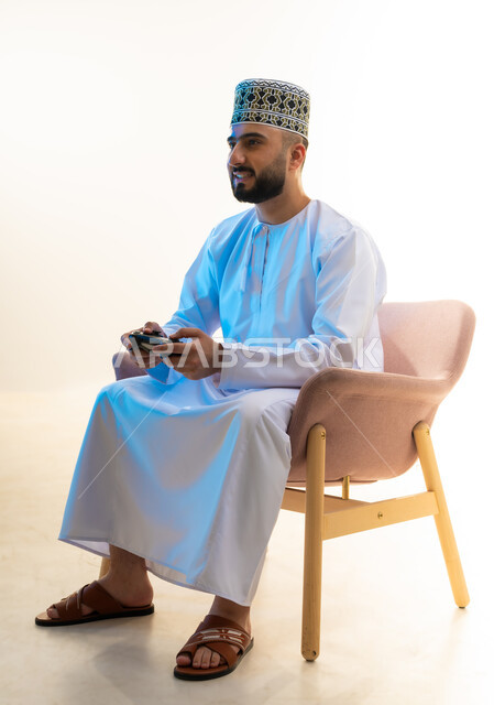 ايماءات السرور والسعادة، التفاعل والاندماج مع ألعاب الفيديو، قضاء وقت ممتع ومسلي، اجهزة وتقنيات الكترونية ترفيهيه حديثة, بورتريه لشاب عربي خليجي عماني يرتدي الدشداشة والكمة يجلس على كرسي مريح يحمل عصا التحكم، خلفية بيضاء
