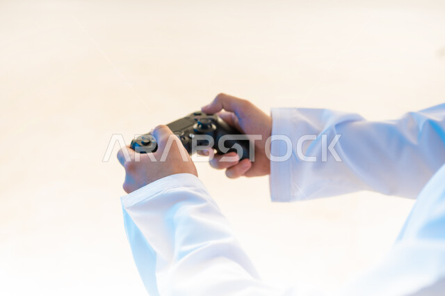 Interacting and merging with video games, having a fun and entertaining time, modern entertainment electronic devices and technologies, close-up portrait from the side of the hands of a young Arab Gulf Omani man wearing a dishdasha holding a joystick, white background