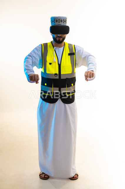 دمج التكنولوجيا بالمجال الهندسي، محاكاة العالم الخيالي، التسلية بالمشاهدة عبر نظارة الواقع الافتراضي، بورتريه لمهندس عربي خليجي عماني يرتدي الدشداشة والكمة وسترة الحماية يضع نظارة الفي ار يمد قبضتيه بايماءات الامساك بشيء ما، صورة لكامل الجسم، خلفية بيضاء