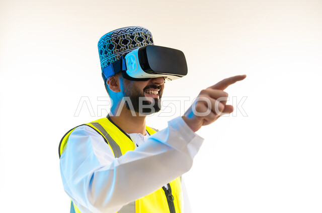 تحديد موقع شيء ما، التسلية بالمشاهدة عبر نظارة الواقع الافتراضي، محاكاة العالم الخيالي، دمج التكنولوجيا بالمجال الهندسي، بورتريه مقرب من الجانب لمهندس عربي خليجي عماني يرتدي الدشداشة والكمة وسترة الحماية يضع نظارة الفي ار يشير باصبع السبابة الى مكان ما، خلفية بيضاء