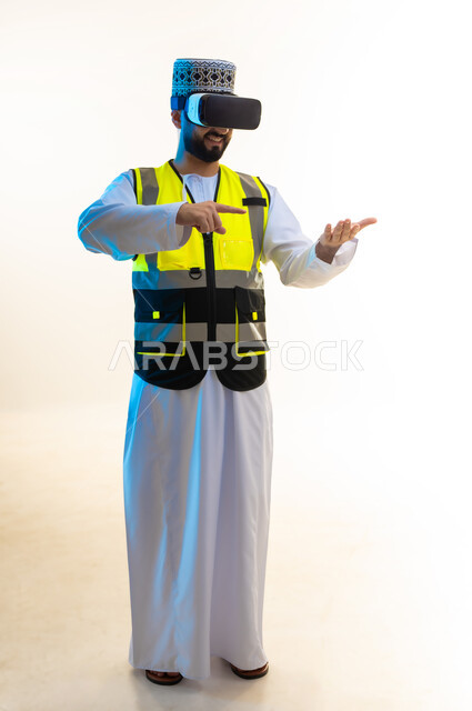 تعابيرحمل عنصر ما، التسلية بالمشاهدة عبر نظارة الواقع الافتراضي، محاكاة العالم الخيالي، دمج التكنولوجيا بالمجال الهندسي، بورتريه لمهندس عربي خليجي عماني يرتدي الدشداشة والكمة وسترة الحماية يضع نظارة الفي ار يشير باصبع السبابة الى يده، صورة لكامل الجسم، خلفية بيضاء