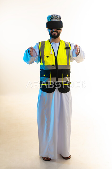 تعابيرحمل عنصر ما، التسلية بالمشاهدة عبر نظارة الواقع الافتراضي، محاكاة العالم الخيالي، دمج التكنولوجيا بالمجال الهندسي، بورتريه لمهندس عربي خليجي عماني يرتدي الدشداشة والكمة وسترة الحماية يضع نظارة الفي ار يرفع يديه بايماءات التحجيم، صورة لكامل الجسم، خلفية بيضاء