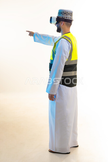 التسلية بالمشاهدة عبر نظارة الواقع الافتراضي، محاكاة العالم الخيالي، دمج التكنولوجيا بالمجال الهندسي، بورتريه من الجانب لمهندس عربي خليجي عماني يرتدي الدشداشة والكمة وسترة الحماية يضع نظارة الفي ار يشير باصبع السبابة لمكان ما، صورة لكامل الجسم، خلفية بيضاء
