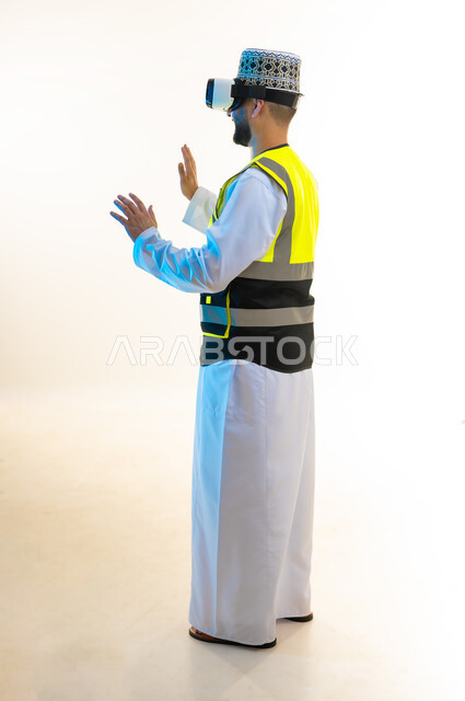 محاكاة العالم الخيالي، دمج التكنولوجيا بالمجال الهندسي، التسلية بالمشاهدة عبر نظارة الواقع الافتراضي، بورتريه من الجانب لمهندس عربي خليجي عماني يرتدي الدشداشة والكمة وسترة الحماية يضع نظارة الفي ار يرفع يديه بايماءات الاندماج، صورة لكامل الجسم، خلفية بيضاء
