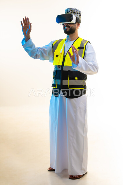 دمج التكنولوجيا بالمجال الهندسي، التسلية بالمشاهدة عبر نظارة الواقع الافتراضي، محاكاة العالم الخيالي، بورتريه لمهندس عربي خليجي عماني يرتدي الدشداشة والكمة وسترة الحماية يضع نظارة الفي ار يرفع يديه بايماءات الاندماج، صورة لكامل الجسم، خلفية بيضاء