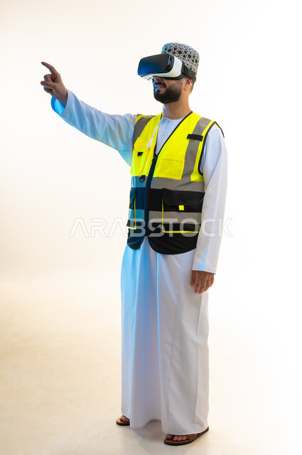 تحديد موقع شيء ما، التسلية بالمشاهدة عبر نظارة الواقع الافتراضي، محاكاة العالم الخيالي، دمج التكنولوجيا بالمجال الهندسي، بورتريه لمهندس عربي خليجي عماني يرتدي الدشداشة والكمة وسترة الحماية يضع نظارة الفي ار يشير باصبع السبابة الى مكان ما، صورة لكامل الجسم، خلفية بيضاء