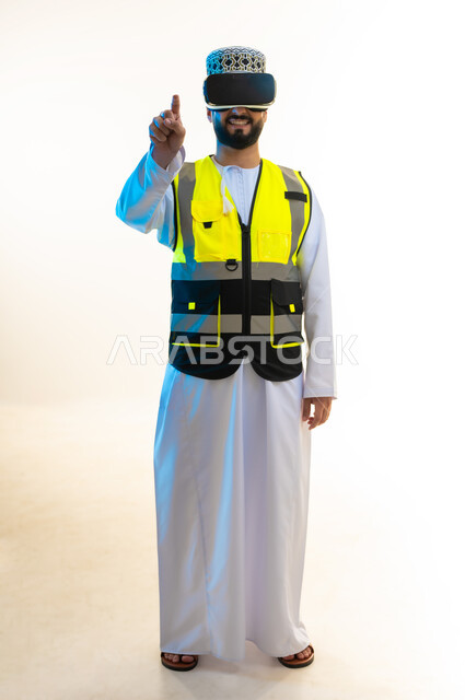 التسلية بالمشاهدة عبر نظارة الواقع الافتراضي، محاكاة العالم الخيالي، دمج التكنولوجيا بالمجال الهندسي، بورتريه لمهندس عربي خليجي عماني يرتدي الدشداشة والكمة وسترة الحماية يضع نظارة الفي ار يشير باصبع السبابة الى الكاميرا، صورة لكامل الجسم، خلفية بيضاء