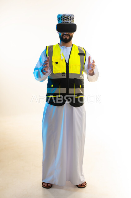 تعابير حمل عنصر ما، التسلية بالمشاهدة عبر نظارة الواقع الافتراضي، محاكاة العالم الخيالي، دمج التكنولوجيا بالمجال الهندسي، بورتريه لمهندس عربي خليجي عماني يرتدي الدشداشة والكمة وسترة الحماية يضع نظارة الفي ار يرفع يديه بايماءات التحجيم، صورة لكامل الجسم، خلفية بيضاء