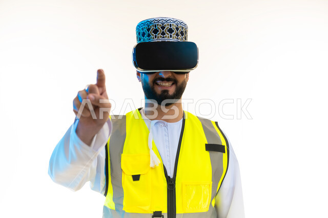 تحديد موقع شيء ما، التسلية بالمشاهدة عبر نظارة الواقع الافتراضي، محاكاة العالم الخيالي، دمج التكنولوجيا بالمجال الهندسي، بورتريه مقرب لمهندس عربي خليجي عماني يرتدي الدشداشة والكمة وسترة الحماية يضع نظارة الفي ار يشير باصبع السبابة الى الكاميرا، خلفية بيضاء