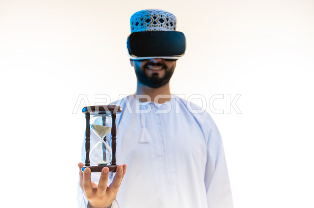 مراقبة وقياس الوقت، محاكاة العالم الخيالي، التسلية بالمشاهدة عبر نظارة الواقع الافتراضي، بورتريه مقرب لرجل عربي خليجي عماني يرتدي الدشداشة والكمة وسترة الحماية يضع نظارة الفي ار يحمل بيده ساعة رملية (البنكام)، خلفية بيضاء