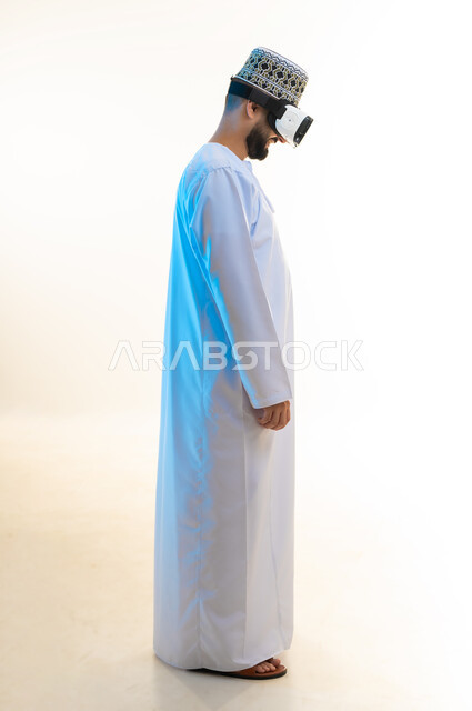 Integration and simulation of the imaginary world, fun and entertainment by watching through virtual reality glasses, technical and technological development, side portrait of an Arab Gulf Omani young man wearing a dishdasha and a kimono looking through 3D glasses (VR), full body image, white background