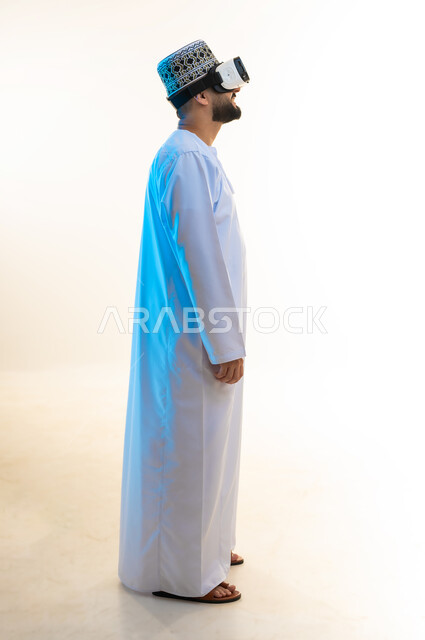 المتعة والتسلية بالمشاهدة عبر نظارة الواقع الافتراضي، التطور التقني والتكنولوجي, بورتريه من الجانب لشاب عربي خليجي عماني يرتدي الدشداشة والكمه ينظر من خلال نظارات ثلاثية الأبعاد (الفي ار), الاندماج ومحاكاة العالم الخيالي، صورة الجسم بالكامل، خلفية بيضاء