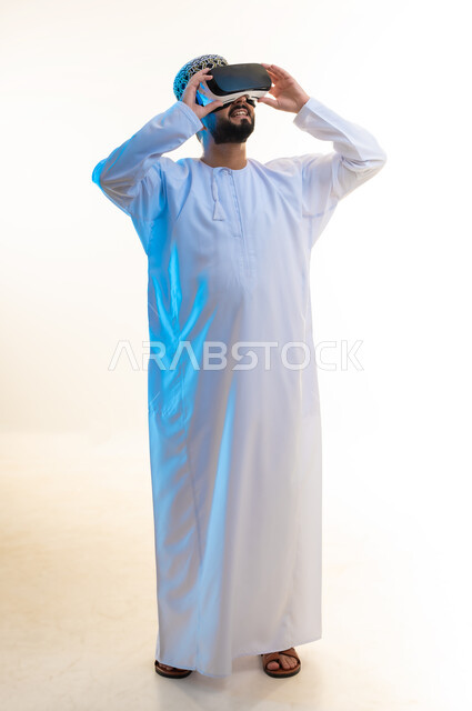 المتعة والتسلية بالمشاهدة عبر نظارة الواقع الافتراضي، التطور التقني والتكنولوجي, بورتريه لشاب عربي خليجي عماني يرتدي الدشداشة والكمه ينظر من خلال نظارات ثلاثية الأبعاد (الفي ار), الاندماج ومحاكاة العالم الخيالي، صورة الجسم بالكامل، خلفية بيضاء