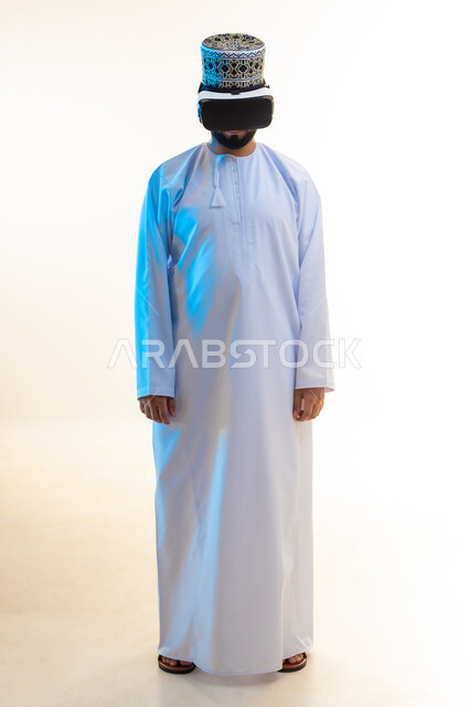 Technical and technological development, fun and entertainment by watching through virtual reality glasses, portrait of a young Arab Gulf Omani man wearing a dishdasha and a kimono looking through 3D glasses (VR), integration and simulation of the imaginary world, full body image, white background