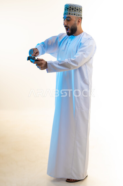 Having a fun and entertaining time, modern entertainment devices and electronic technologies, interacting and merging with video games, portrait of a young Arab Gulf Omani man wearing a dishdasha and a turban pressing the joystick buttons with gestures of surprise, full body image, white background