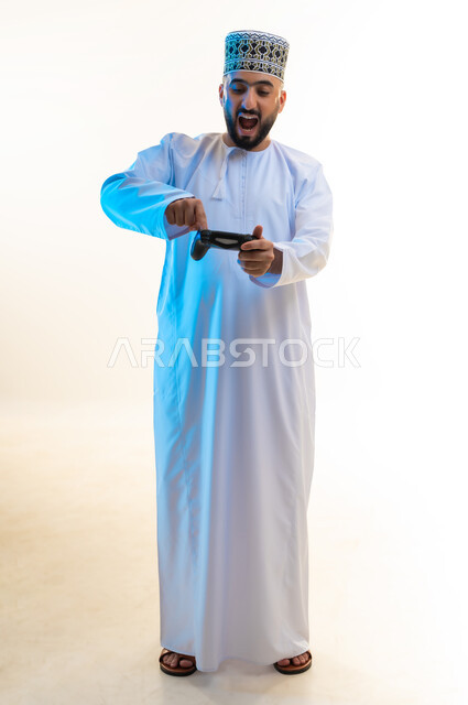 قضاء وقت ممتع ومسلي، اجهزة وتقنيات الكترونية ترفيهيه حديثة, التفاعل والاندماج مع ألعاب الفيديو، بورتريه لشاب عربي خليجي عماني يرتدي الدشداشة والكمة يقوم بالضغط على ازرار عصا التحكم بايماءات الدهشة، صورة لكامل الجسم، خلفية بيضاء