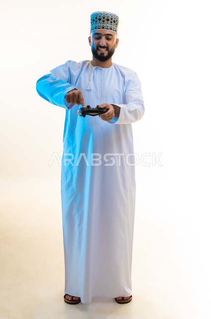 اجهزة وتقنيات الكترونية ترفيهيه حديثة, قضاء وقت ممتع ومسلي، التفاعل والاندماج مع ألعاب الفيديو، بورتريه لشاب عربي خليجي عماني يرتدي الدشداشة والكمة يقوم بالضغط على ازرار عصا التحكم، صورة لكامل الجسم، خلفية بيضاء